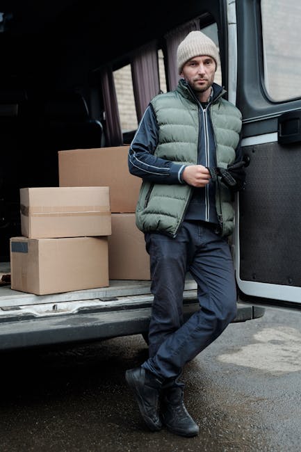 A man wearing a white t-shirt and jeans is outdoors near a residential house, standing on the pavement beside a clothes or furniture removal van with its rear doors open. He is handling a cardboard box, preparing to load or unload it, with additional cardboard boxes stacked on the ground nearby. The van is positioned on the street close to the property's driveway, and the background features a white house with a pitched roof, a wooden fence, and green shrubs and trees. The scene captures a typical home relocation process involving packing and furniture transport to or from a property, with the presence of packing materials such as cardboard boxes and the open van indicating active loading or unloading. Man with Van Uxbridge offers professional removals and transit services, including packing, storage, and home moves, suited for students and residents in the Uxbridge UB8 area.