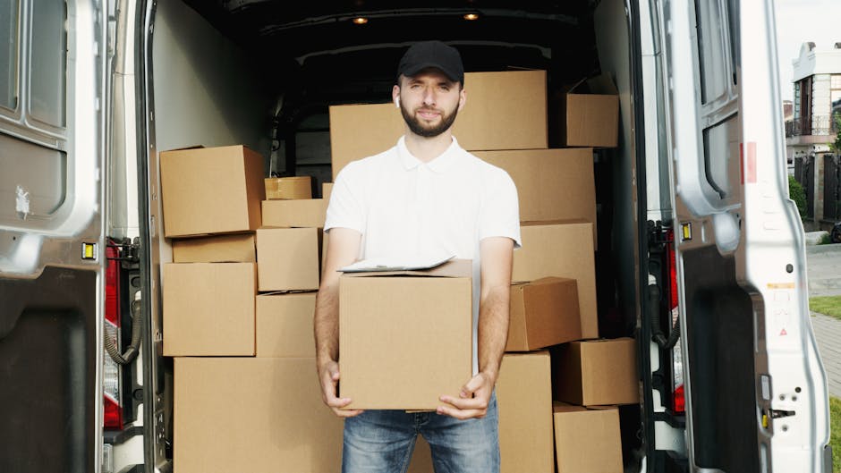 A smiling young man with long, wavy blond hair and a black and white patterned headband is sitting in the driver's seat of a white moving van with the logo 'MOVING COMPANY LOCAL & LONG DISTANCE' print